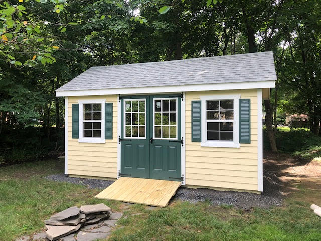 A cream-colored cottage vinyl shed with green shutters and doors and white trim, with light-gray asphalt roofing tiles.